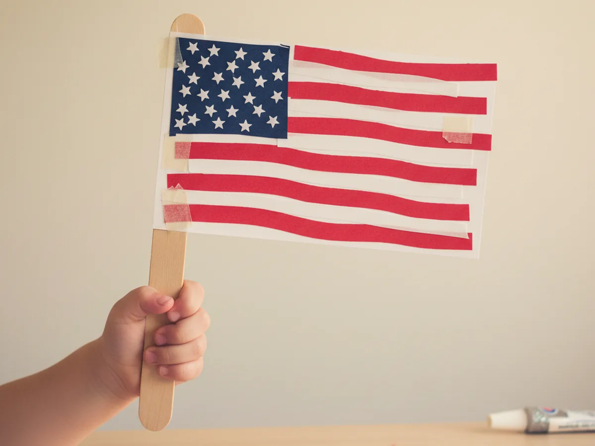 A finished handmade American craft paper flag taped to a jumbo wooden popsicle stick handle with red and white stripes, a navy blue rectangle in the top left filled with small white star stickers, held up in a child's hand against a light wall background