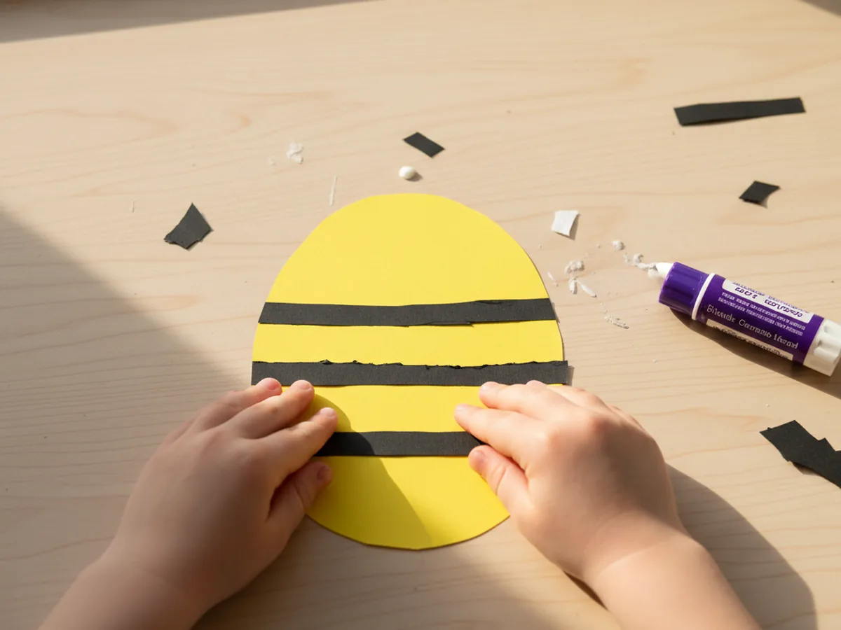 A child gluing three thin black paper stripes across the yellow oval body of a paper bee craft