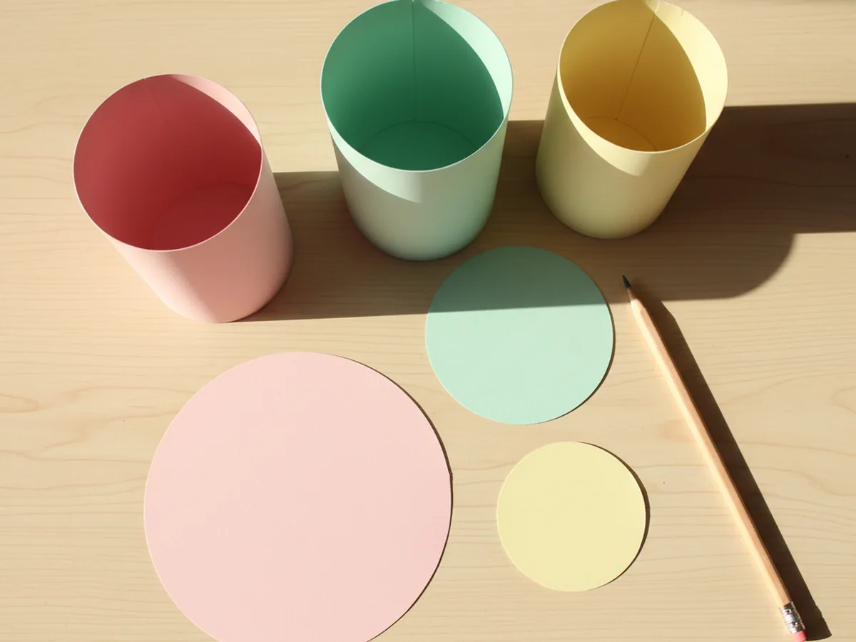 Three flat pastel cardstock circles in pink mint and yellow cut to slightly larger than each cake cylinder, lying on a light wood craft table next to the three rolled paper tiers and a pencil