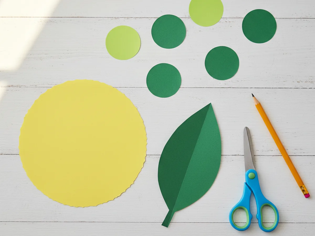 A larger yellow construction paper circle for the caterpillar head and a hand-cut dark green paper leaf shape laid out beside the six green body circles on a white craft table