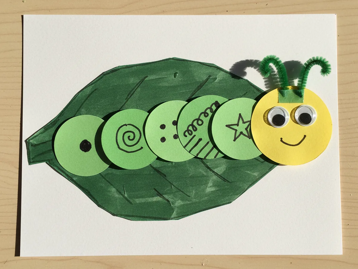 Finished caterpillar paper craft made from six green construction paper circles, a yellow head with googly eyes, two pipe cleaner antennae, and a green leaf on white cardstock