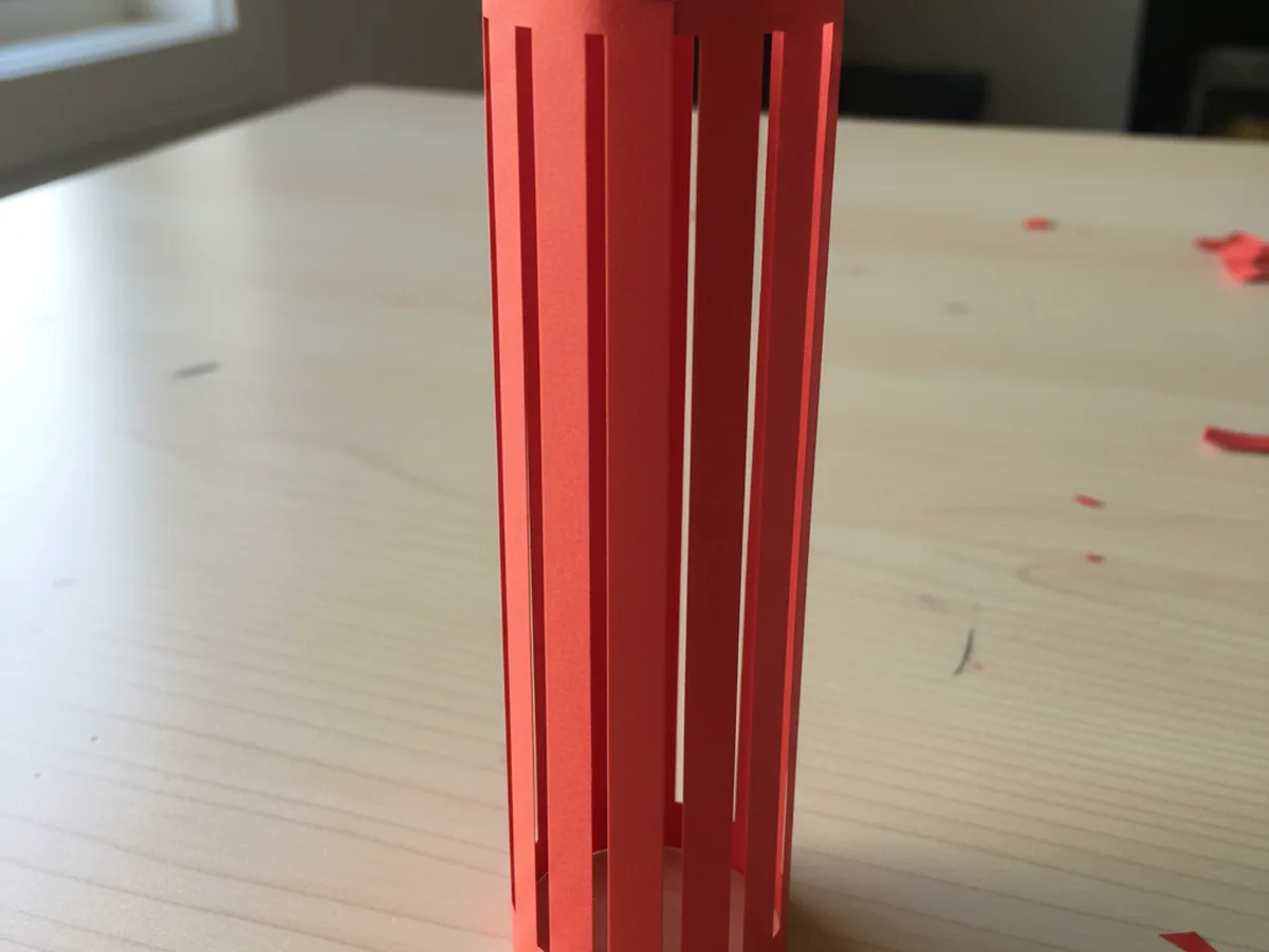 The unfolded red construction paper now rolled into a tall vertical cylinder with the cut slits bowing outward in the middle, the two short ends joined by a small staple, standing upright on a white craft table