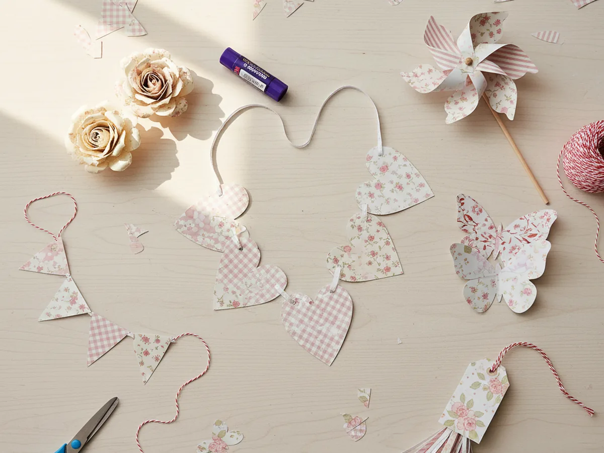 A flat lay of finished craft patterned paper projects including a heart garland, pinwheel, butterflies, bunting, and rolled paper roses on a light wooden craft table