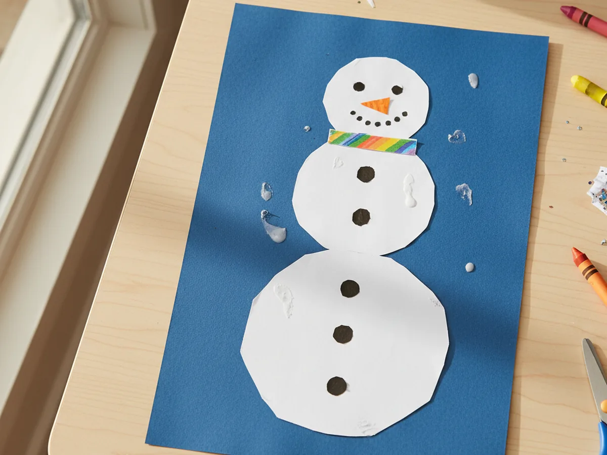 Stacked white construction paper snowman with orange paper nose and a colorful paper scarf on a blue background