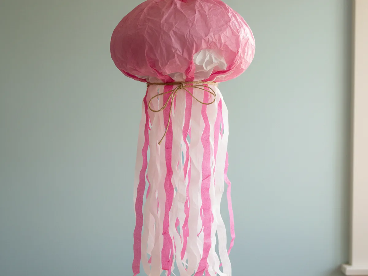 Handmade pink crepe paper jellyfish with a round body and long wavy streamer tentacles hanging from a ceiling
