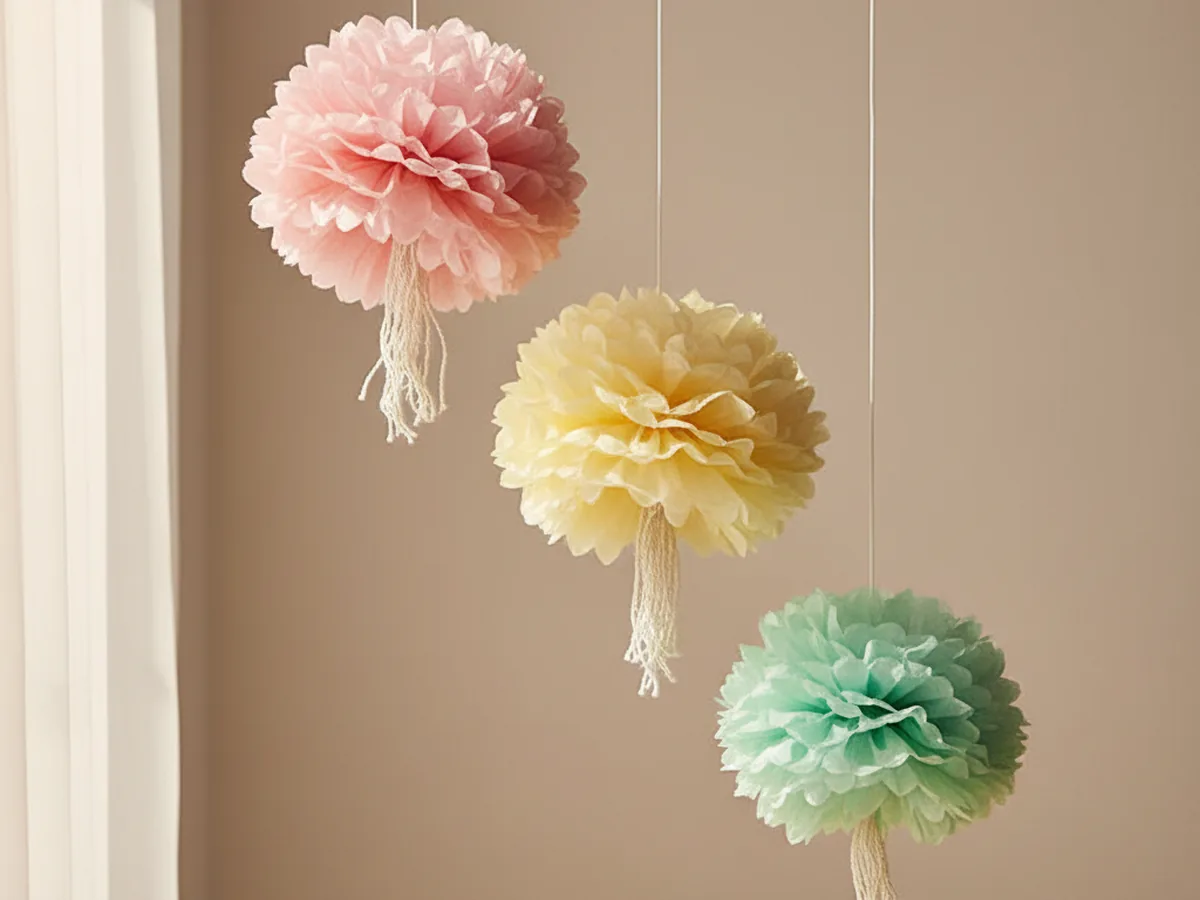 Handmade pastel pink and yellow fluffy crepe paper pom-poms hanging from string above a craft table