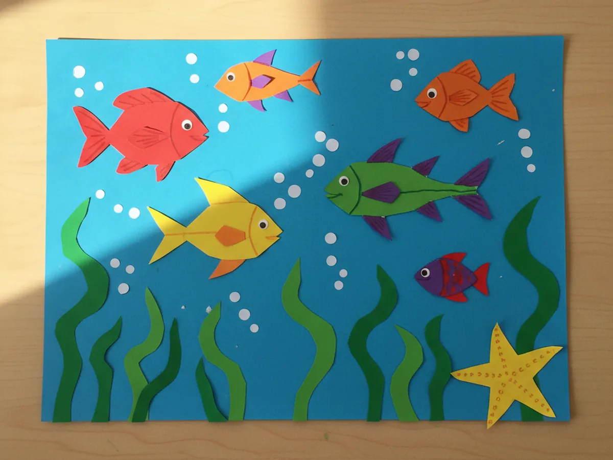 A handmade paper aquarium scene with colorful paper fish, seaweed, and bubbles arranged on a blue background