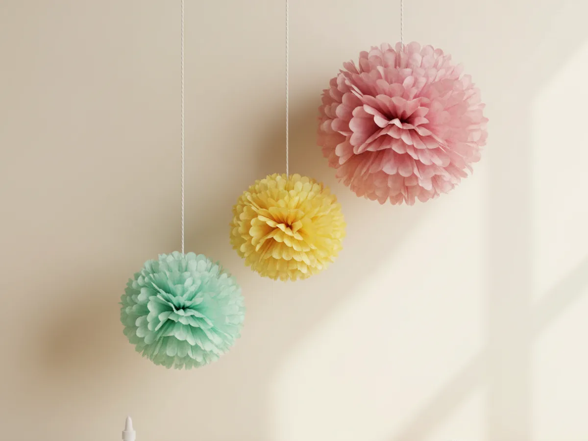 Fluffy handmade tissue paper pom-pom decorations in pink, yellow, and mint green hanging from string on a wall