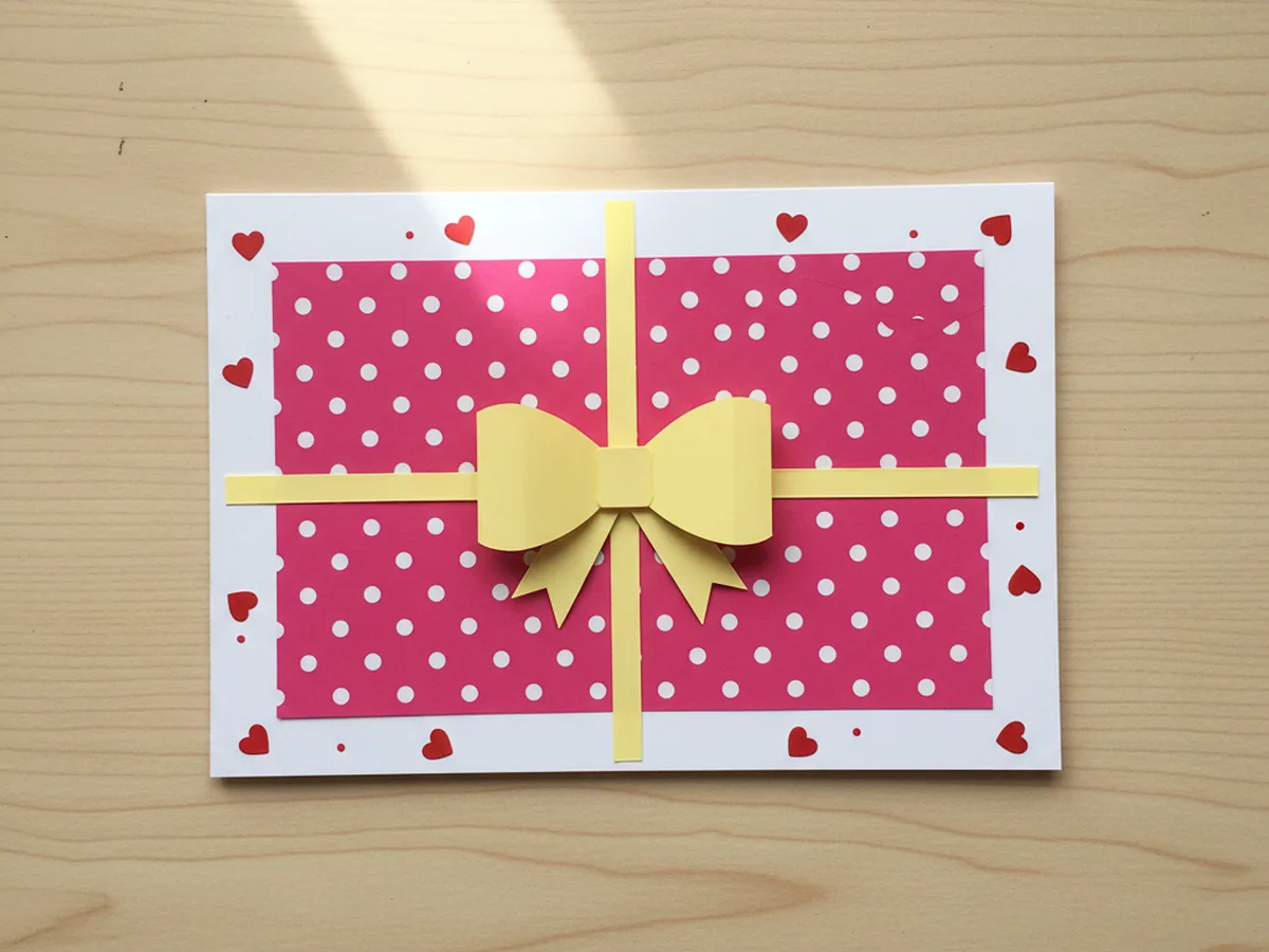 The completed front of the gift paper craft card on a light wood craft table showing the white folded cardstock base with a centered bright pink patterned wrapping rectangle, two thin yellow paper ribbon strips crossing the wrapping, a soft yellow paper bow glued at the center where the ribbon strips meet, and several small red heart stickers and tiny marker dots scattered around the white border