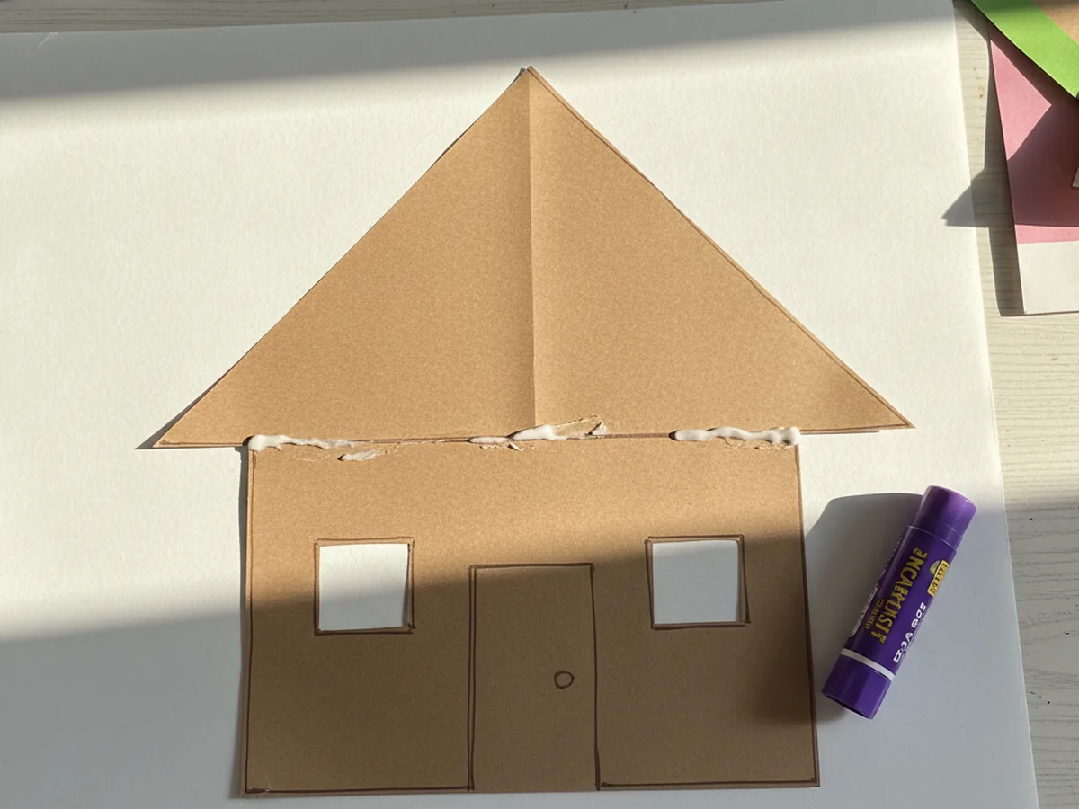 A fully assembled brown paper gingerbread house with the triangle roof glued on top of the square base, a small rectangular door glued in the bottom center, and two square windows on either side of the door, displayed on a white craft table with a glue stick beside it