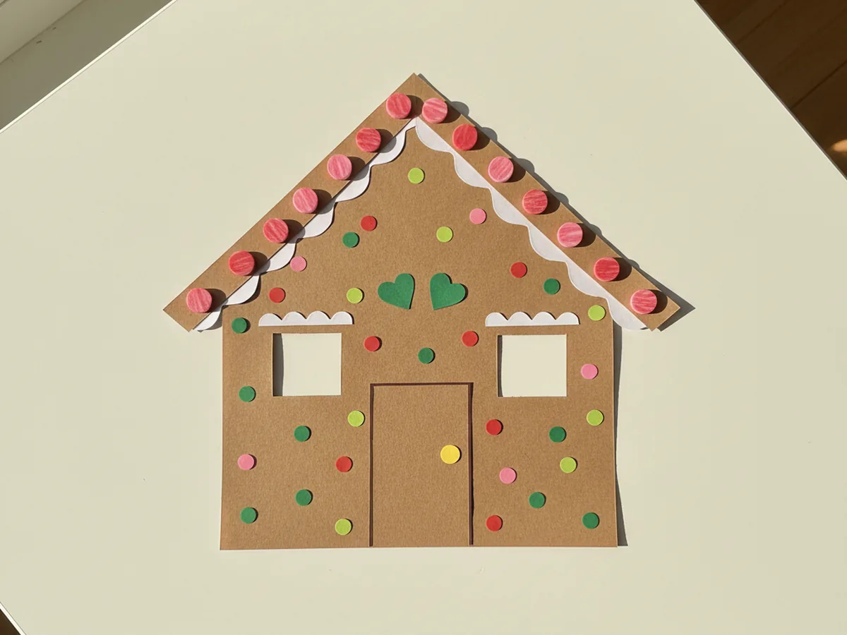 The paper gingerbread house now covered in tiny colorful paper candy details: red and pink dots in a row along the roof like jellybeans, a yellow circle doorknob, small green hearts above the windows, and scattered colored dots all over the walls, displayed on a white craft table