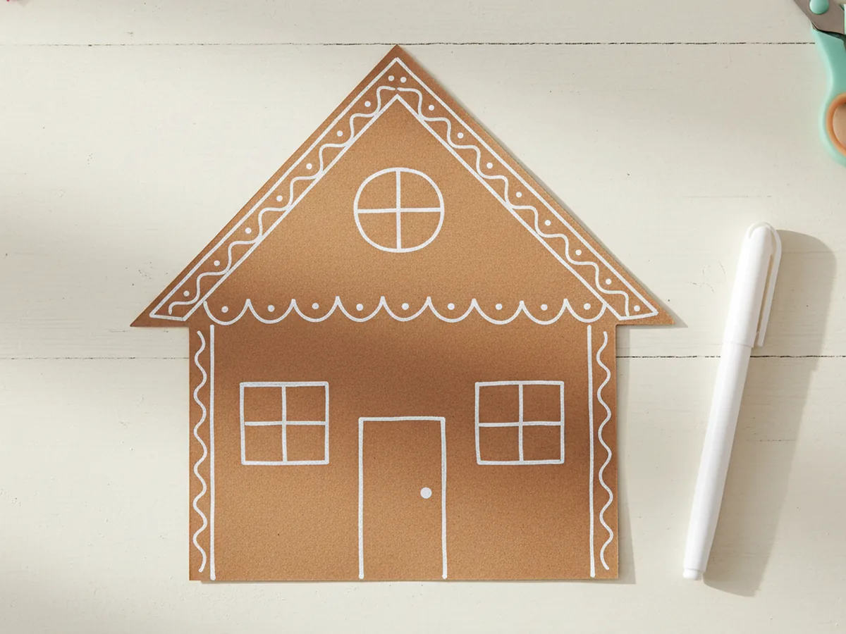 The brown paper gingerbread house now decorated with wavy white icing lines drawn along the roof edge, around the door, around both windows, and along the side edges, looking like piped royal icing, on a white craft table