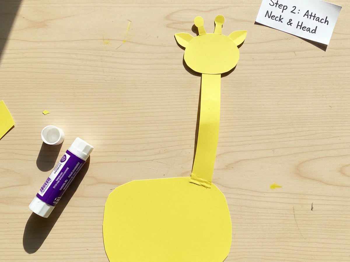 A small rounded yellow paper head with two tiny stubby ossicones glued to the top of the long curved neck of a giraffe paper craft on a craft table