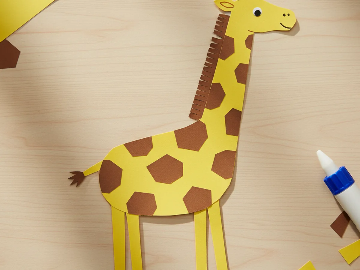 Finished giraffe craft paper made from yellow construction paper with brown spots, a long neck, four tall legs, a brown mane, and small black eyes on a craft table