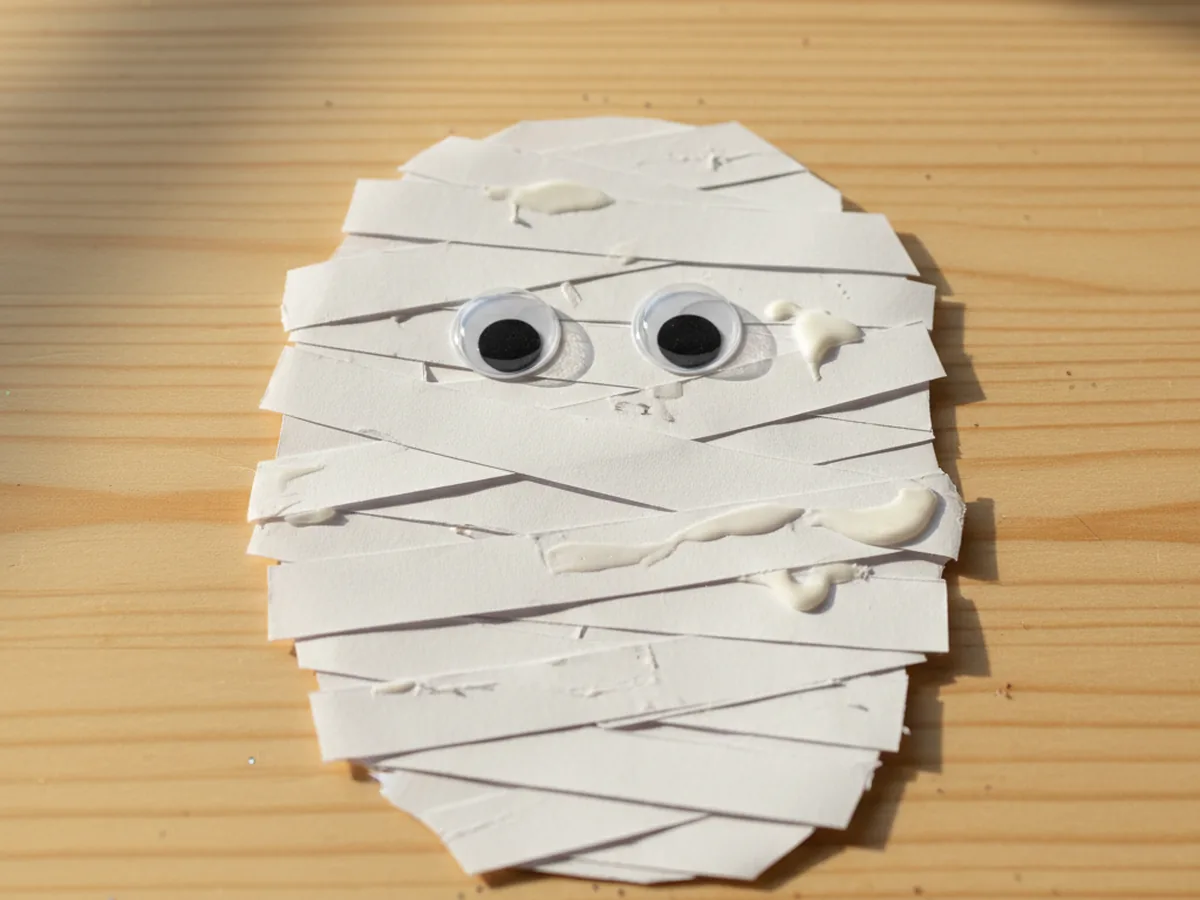 A handmade construction paper mummy with overlapping white paper bandage strips and two googly eyes on a light wood craft table