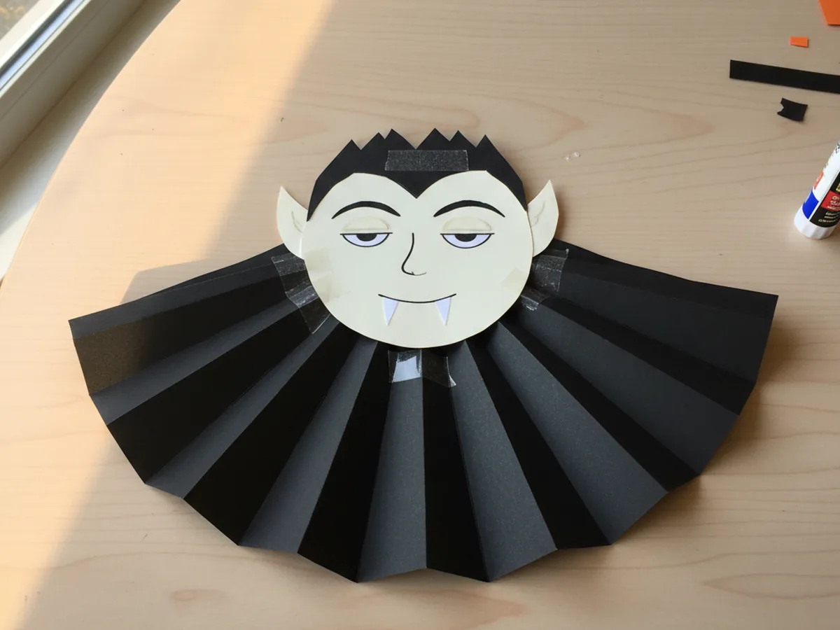 A handmade construction paper vampire with a pale oval face, black widow's peak hair, white paper fangs, and a dramatic black cape