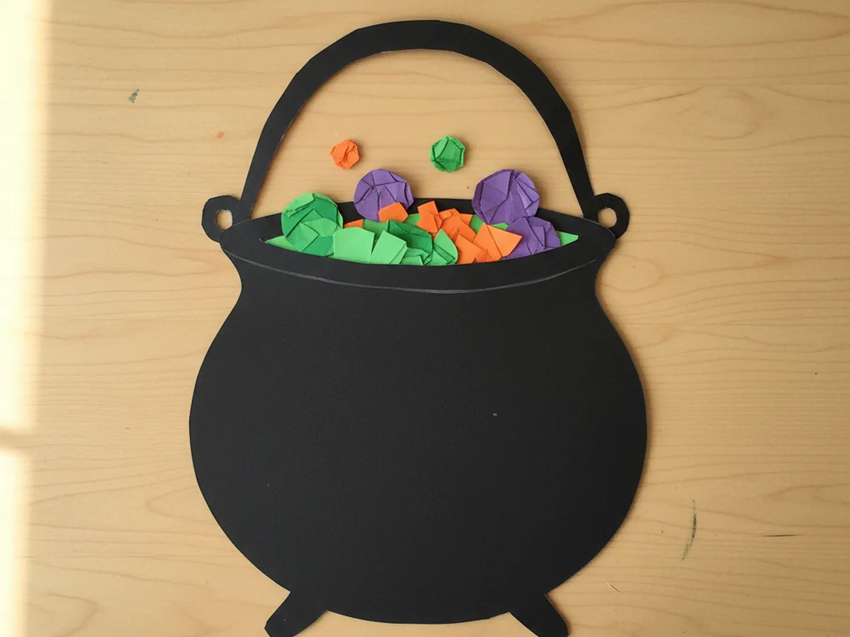 A handmade paper cauldron cut from black construction paper with colorful paper scraps in green and purple inside the top to represent bubbling potion