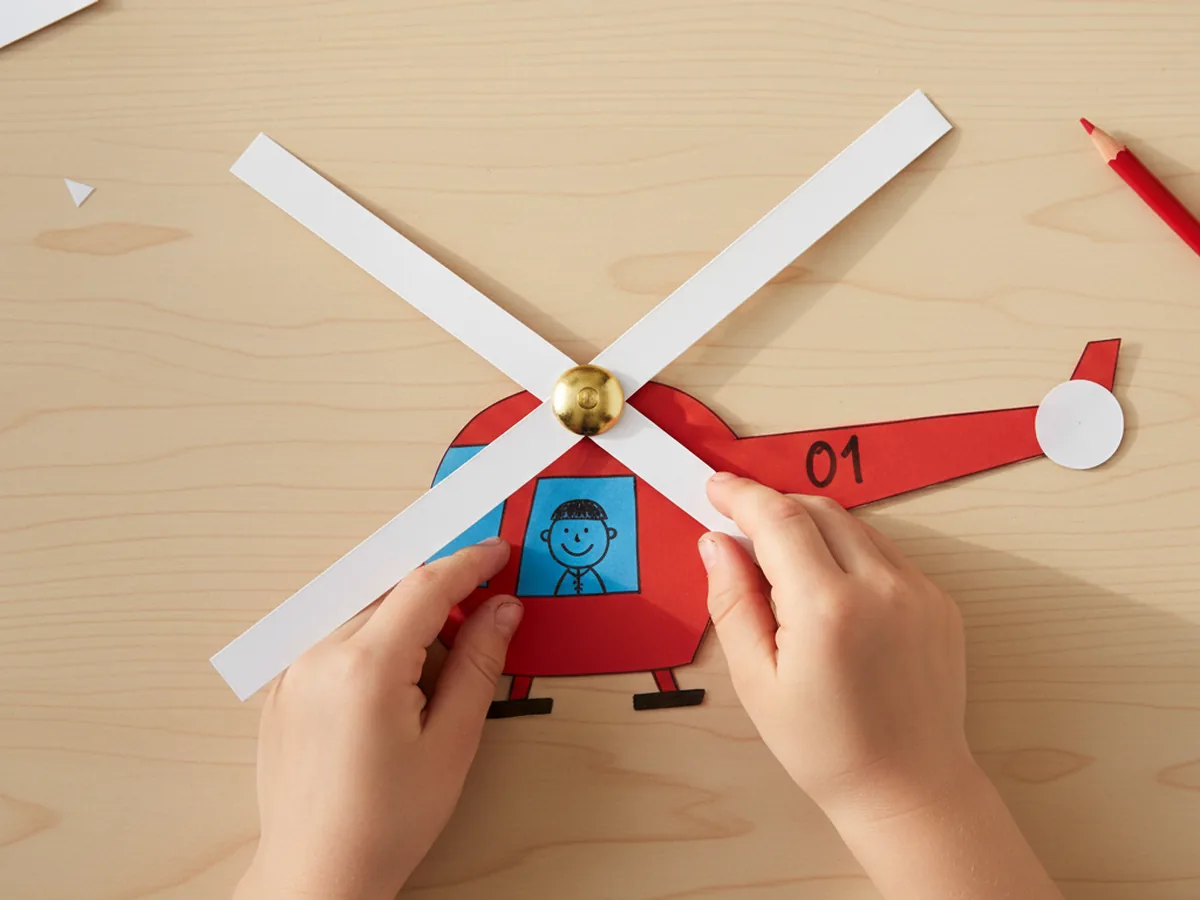 A child's small hands attaching two long white paper rotor blades crossed into an X shape onto the top center of the red helicopter body using a single gold brass brad fastener that is being pushed through both blades and the cardstock body on a light wood craft table