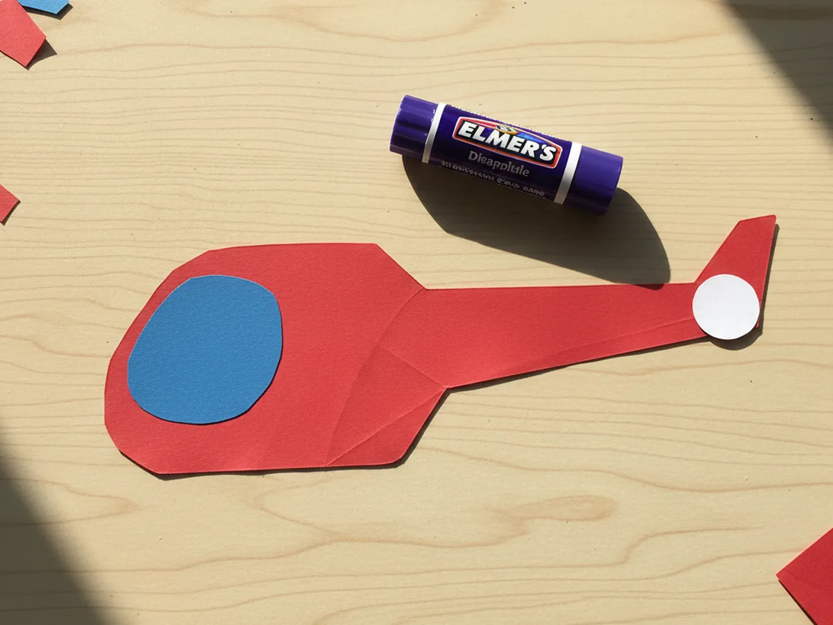 The red helicopter body shape now decorated with a small white circle tail rotor glued at the end of the tail boom and a rounded blue cockpit window glued onto the front cabin area lying flat on a light wood craft table next to a purple glue stick