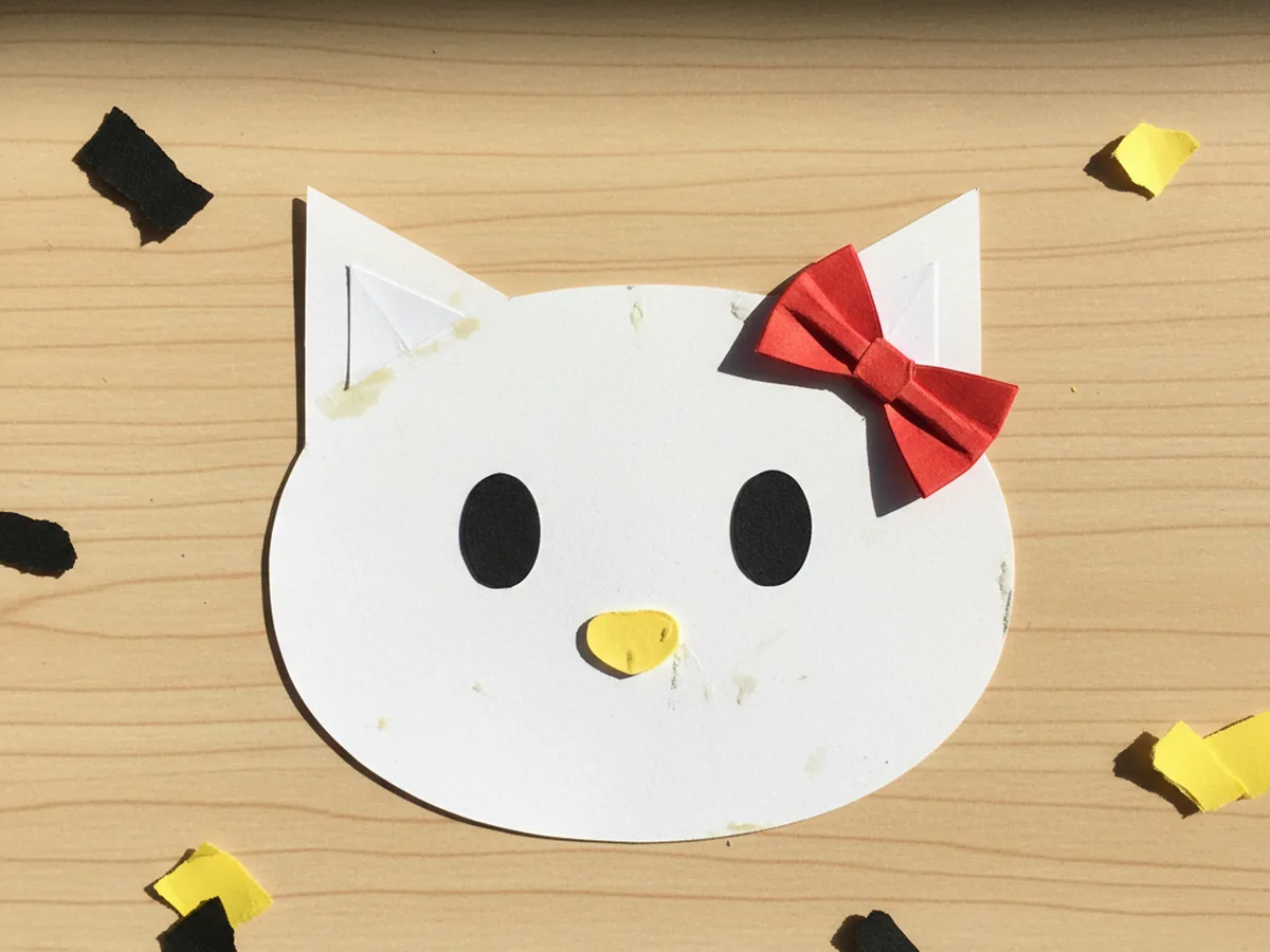 A white paper kitty head with two pointy ears, a red bow, two small black oval eyes, and a small yellow oval nose glued onto the center of the face