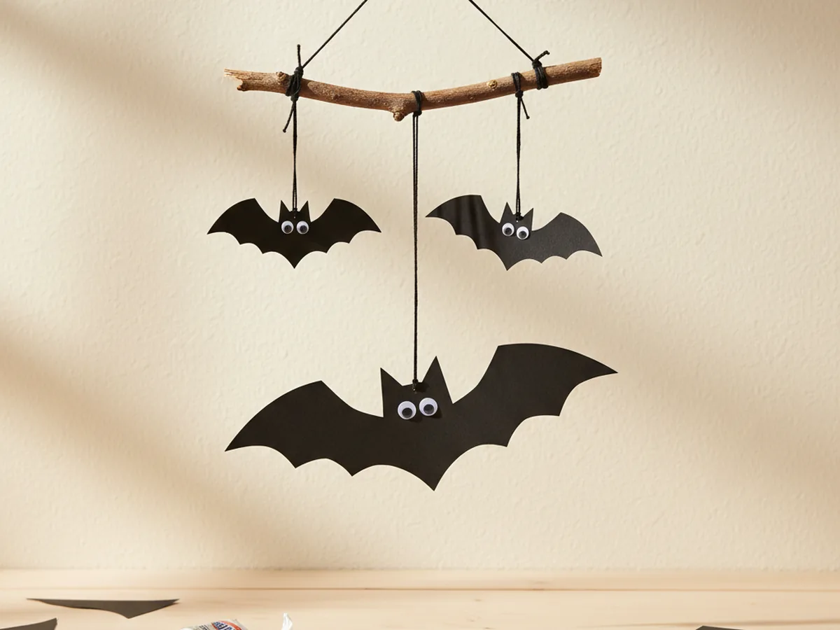A handmade Halloween bat mobile with three black paper bats with googly eyes hanging on strings from a small wooden twig