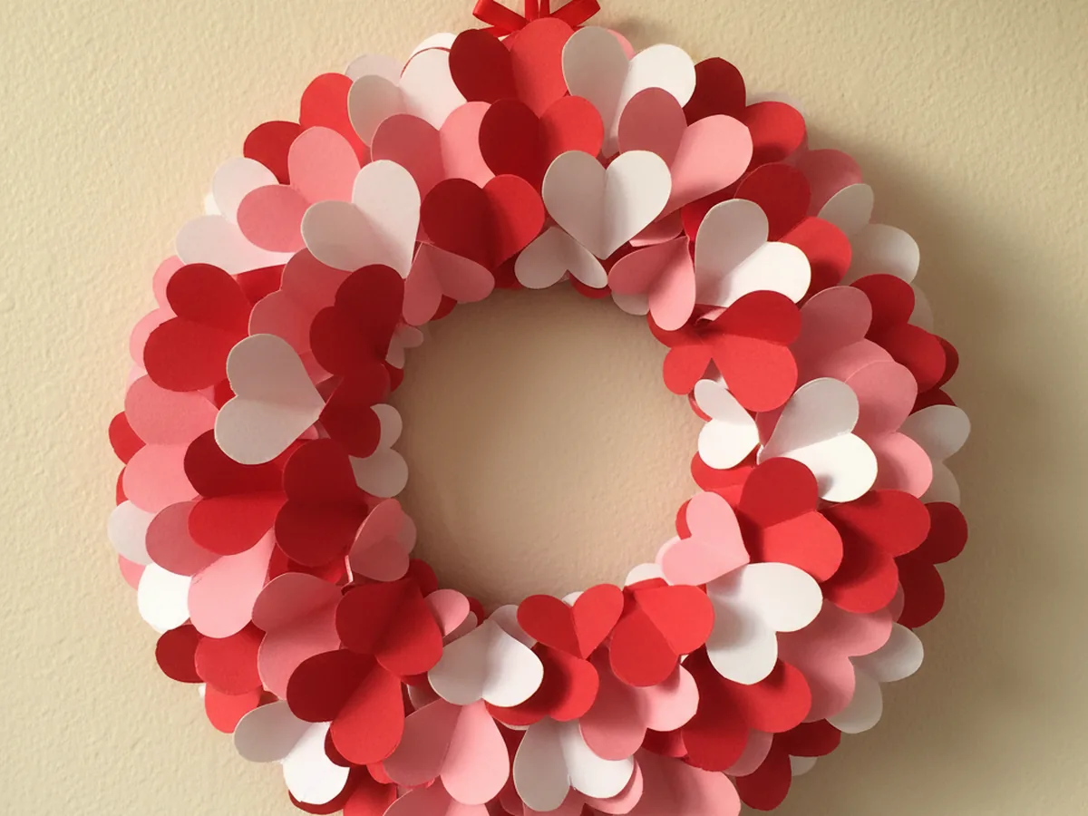 A handmade layered heart paper wreath with pink, red, and white paper hearts glued around a paper plate ring with a ribbon at the top hanging on a wall