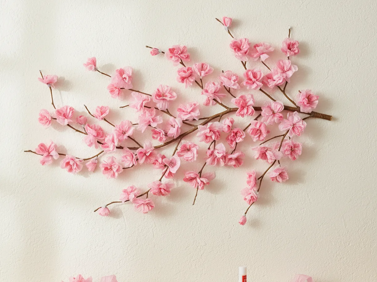 A handmade paper cherry blossom branch with pink tissue paper blossoms attached to a slim brown branch mounted horizontally on a light wall
