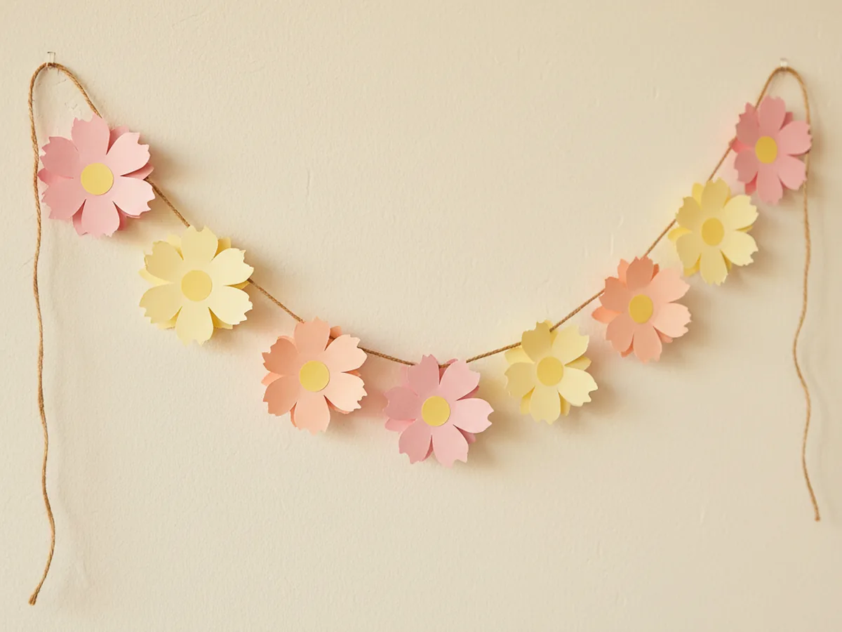 A handmade paper flower wall garland with layered pink, peach, and yellow paper flowers strung on twine and draped against a light wall