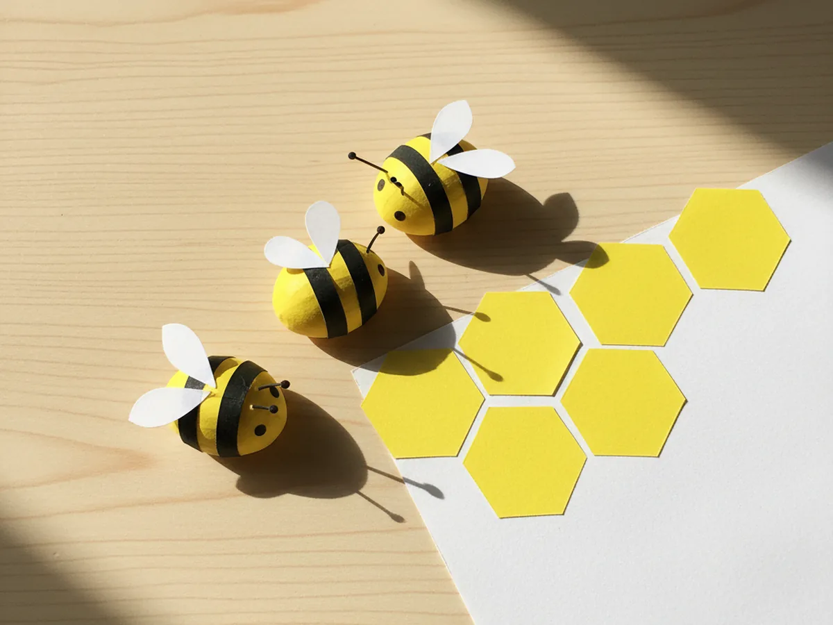 Three small handmade paper bees with white teardrop wings, tiny black eyes, and thin black antennae lying on a craft table next to a yellow honeycomb cluster