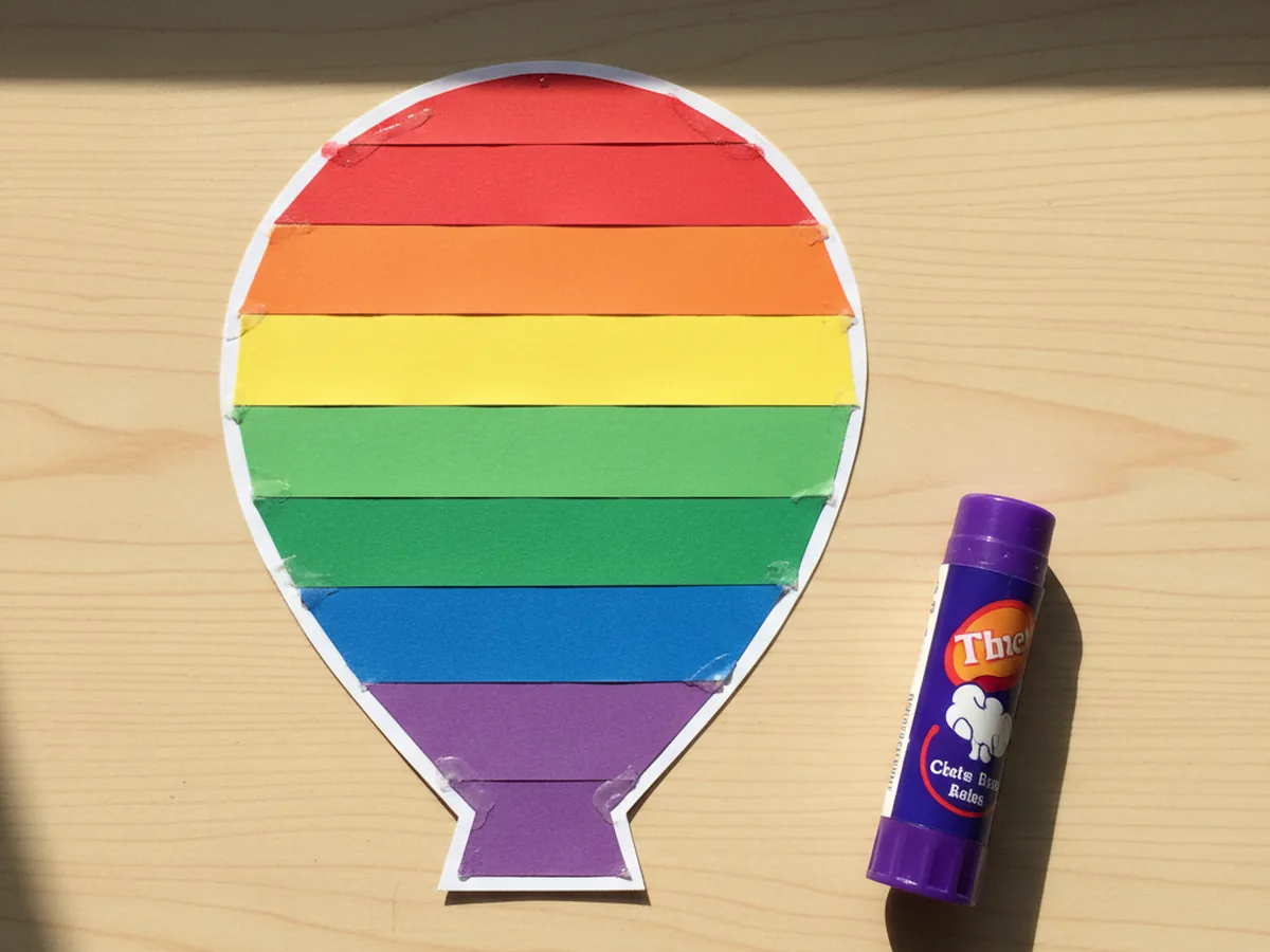 Colorful red, orange, yellow, green, blue, and purple construction paper stripes glued in rainbow order across a rounded cardstock balloon shape for a hot air balloon paper craft