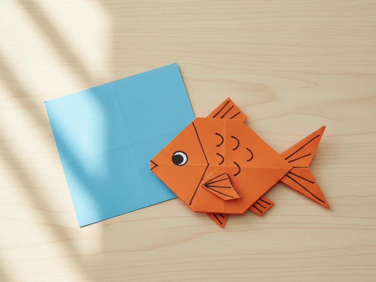 A folded orange origami fish with a marker eye on a light wood table
