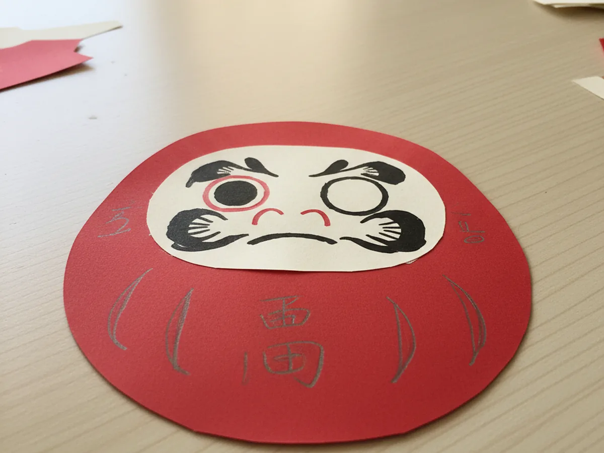 A handmade paper daruma doll cut from red cardstock with a face and one filled eye on a light wood table