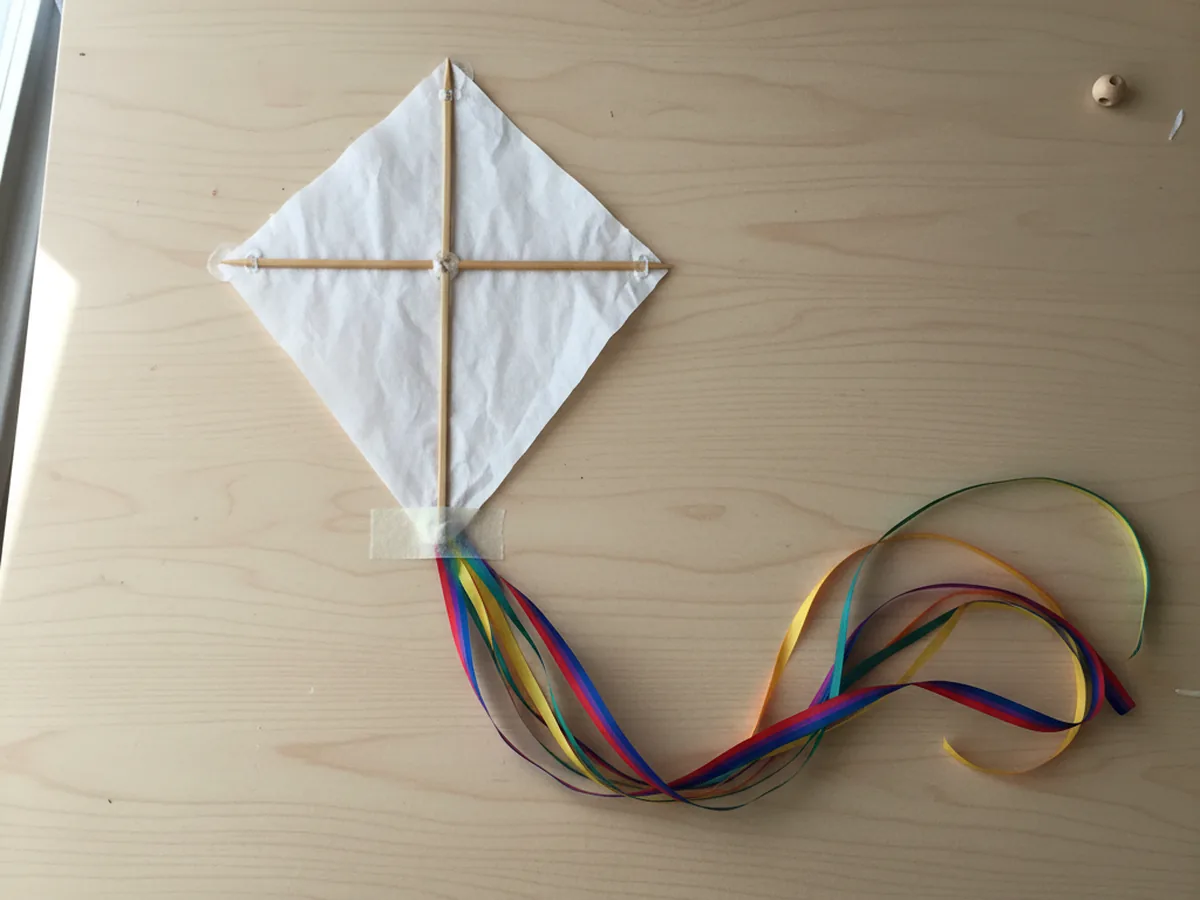 A long rainbow curling ribbon tail taped to the bottom point on the back of a diamond paper kite for the kite craft paper project on a wood craft table