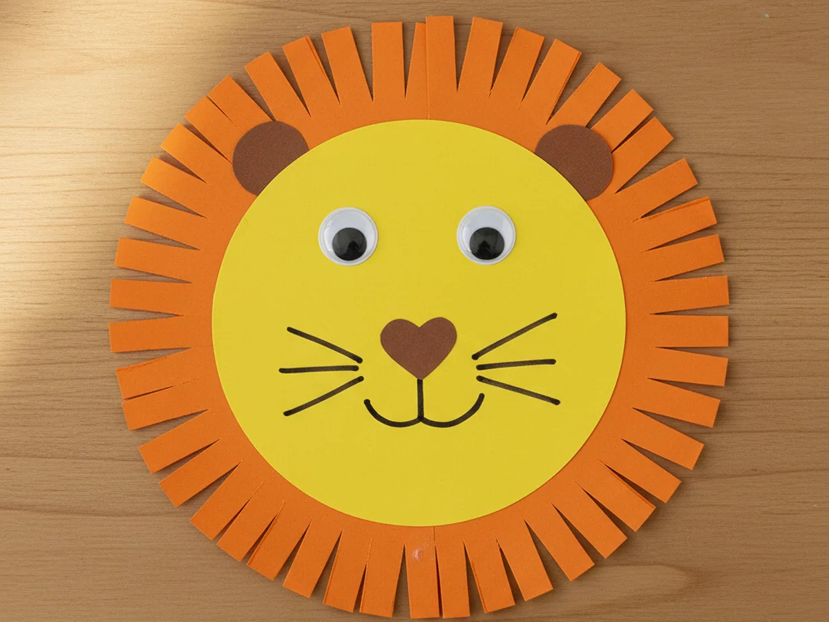 Finished cute lion craft paper with yellow face, fringed orange paper mane, brown ears, and googly eyes on a white craft table