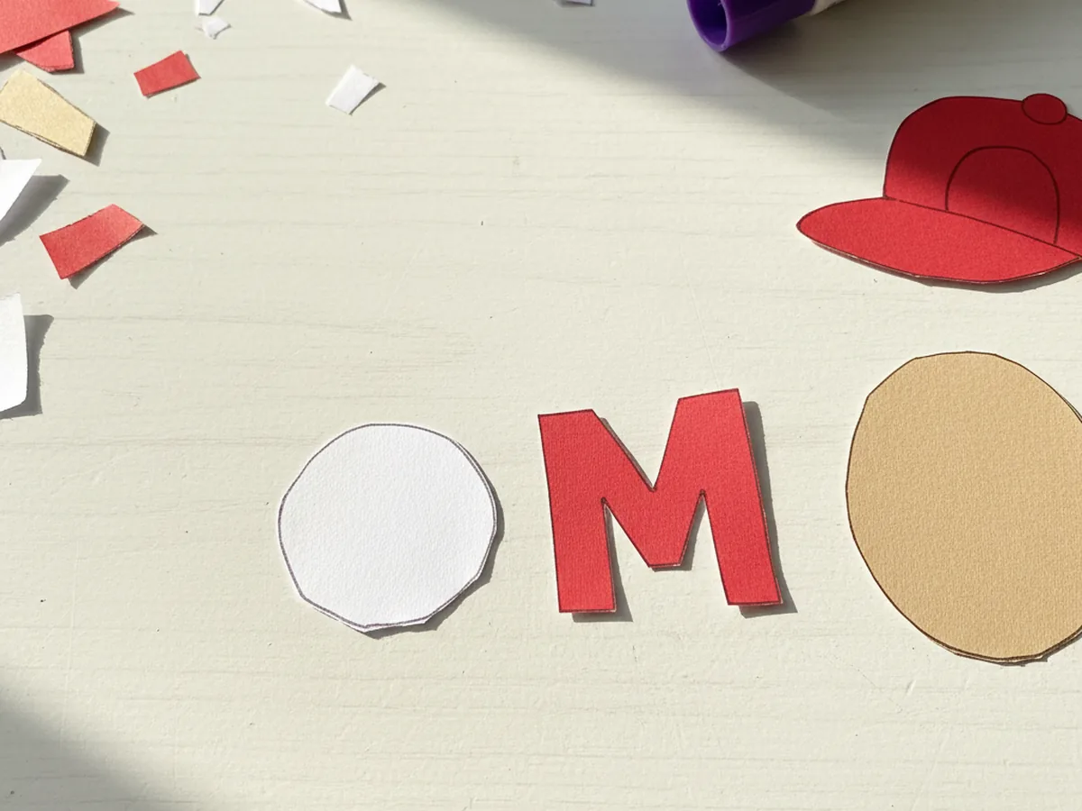 A small white paper circle next to a chunky red letter M cut from construction paper for the Mario paper craft cap logo
