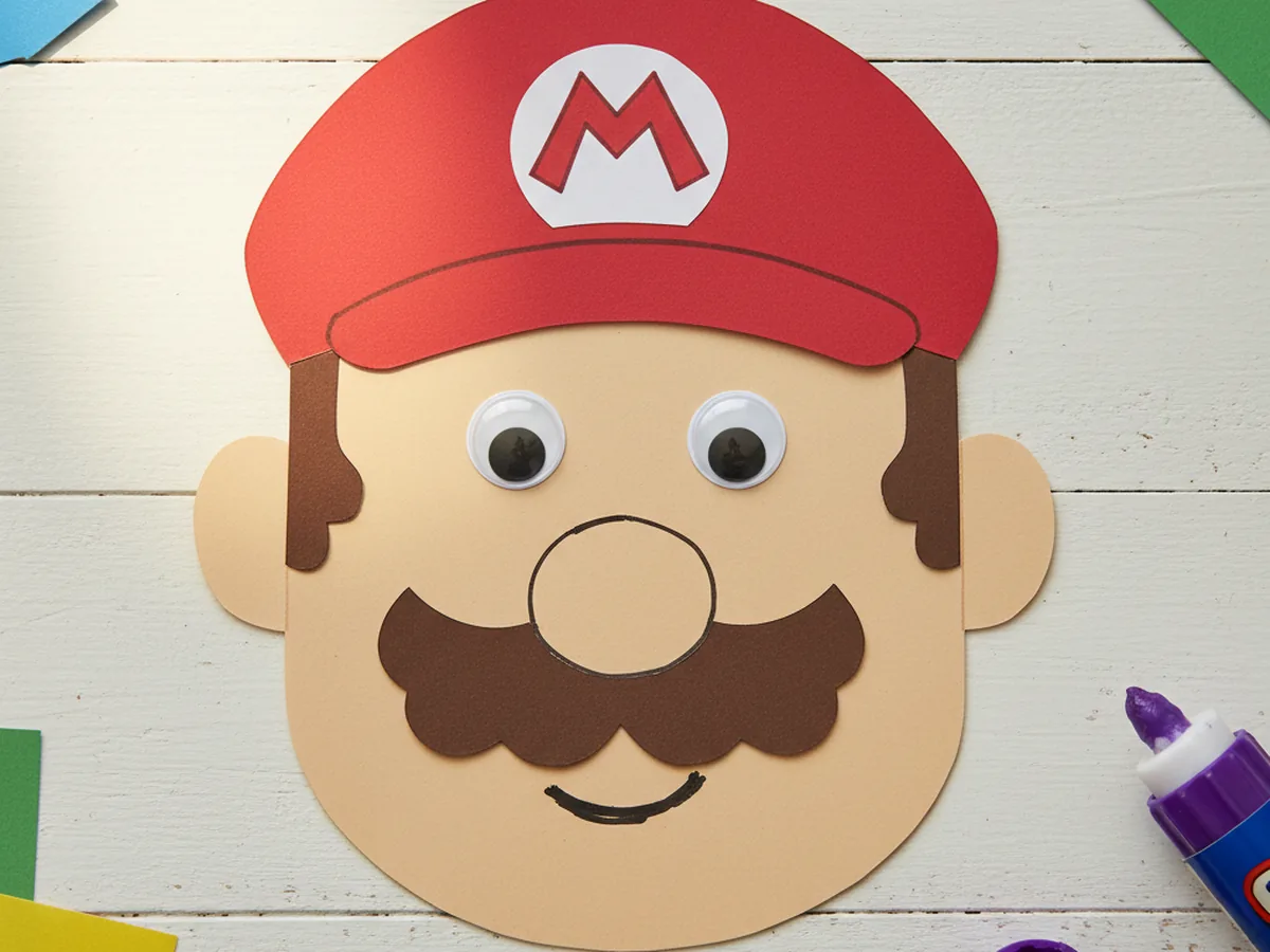 Finished cute Mario paper craft with red cap, white circle and red M logo, tan face, brown mustache, and googly eyes on a white craft table