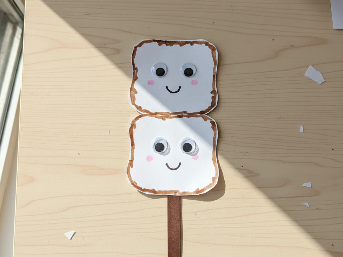 A finished cheerful marshmallow paper craft with two stacked white marshmallows with googly eyes and tiny smiles, lightly toasted edges, on a brown paper roasting stick
