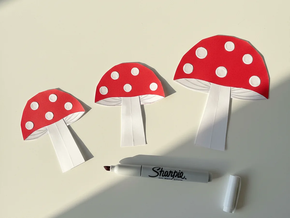 Three tiny paper toadstool mushrooms with red caps freshly decorated with five or six small round white paint marker dots scattered across each cap, lying on a white craft table with an open white paint marker beside them