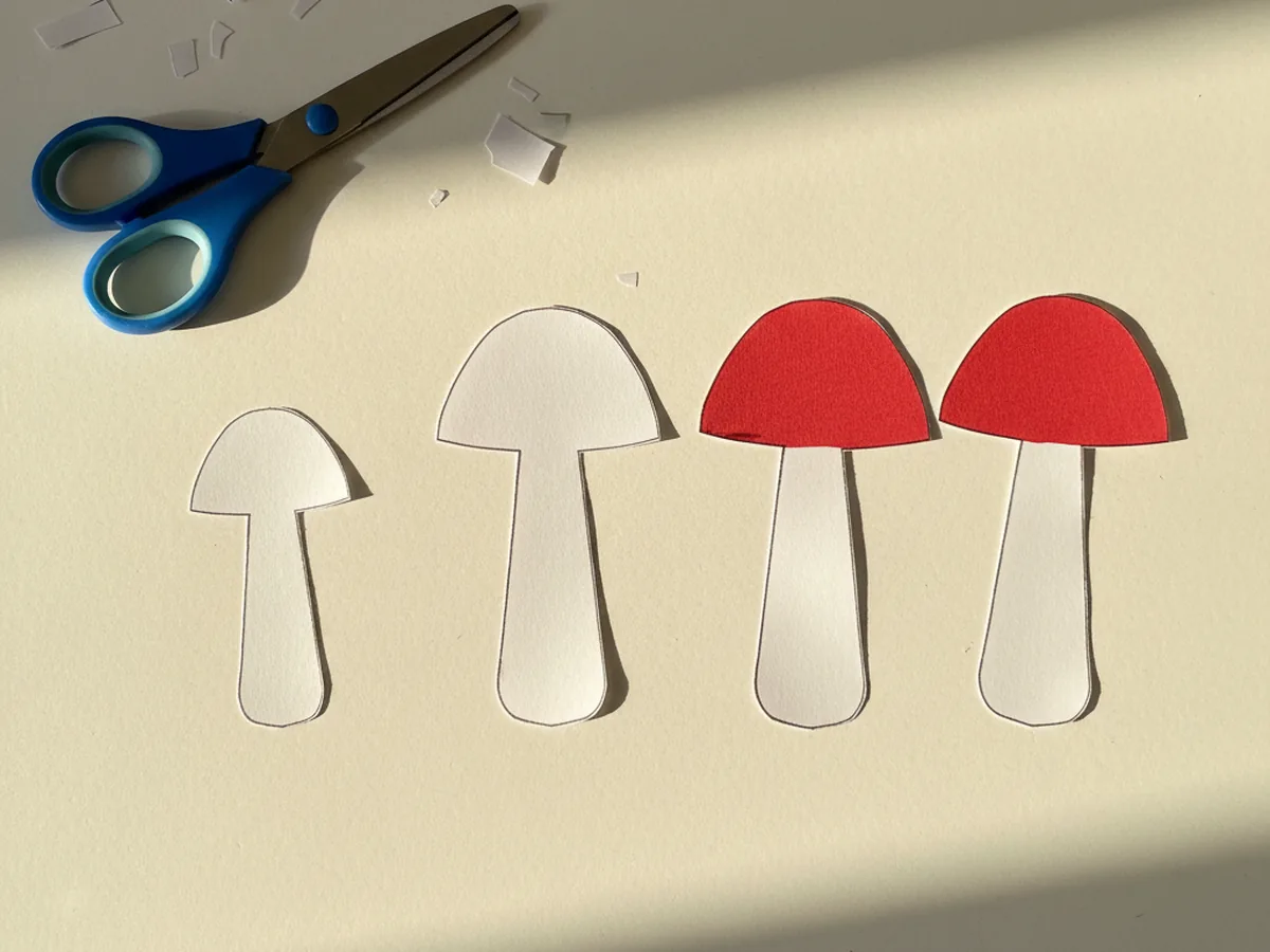 Three small white construction paper mushroom stem shapes in short, medium, and tall sizes with rounded bottom corners, just cut out and lying next to three tiny red mushroom cap dome shapes on a white craft table