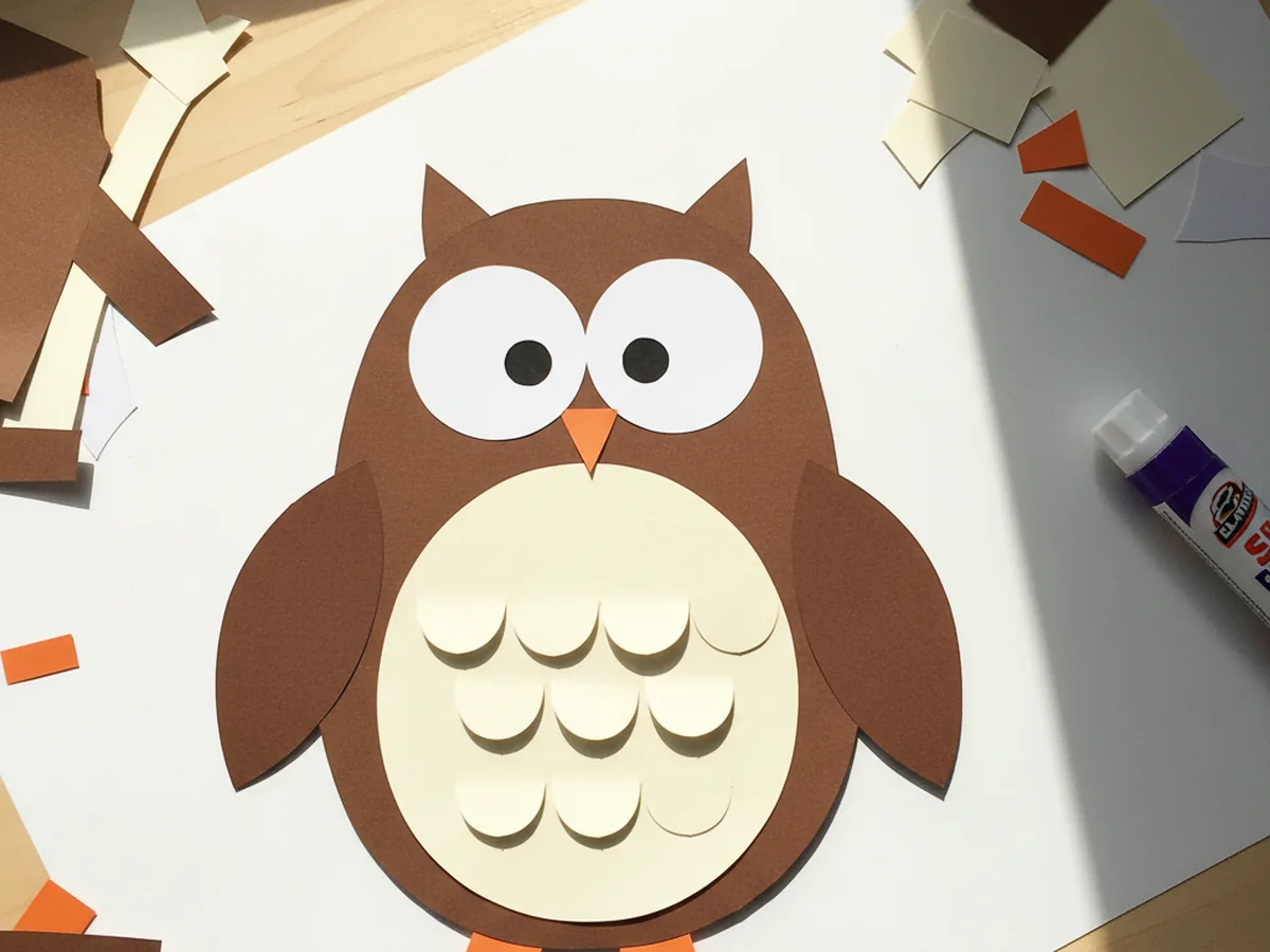 Finished brown owl paper craft with large white eyes, black pupils, an orange triangle beak, brown side wings, scalloped cream feathers across the chest, and small orange feet on a white craft table