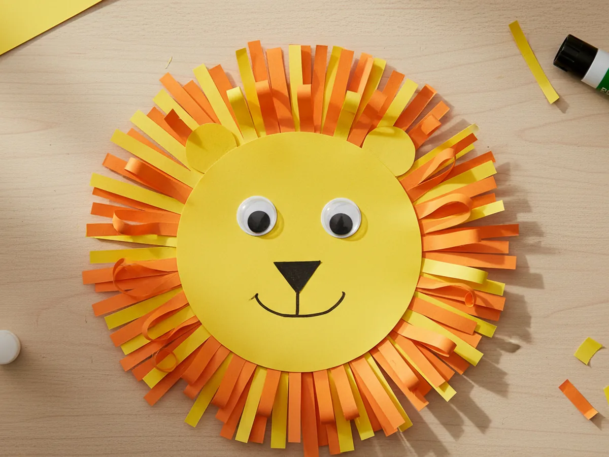 A handmade paper lion with a yellow face and an orange paper strip mane on a craft table