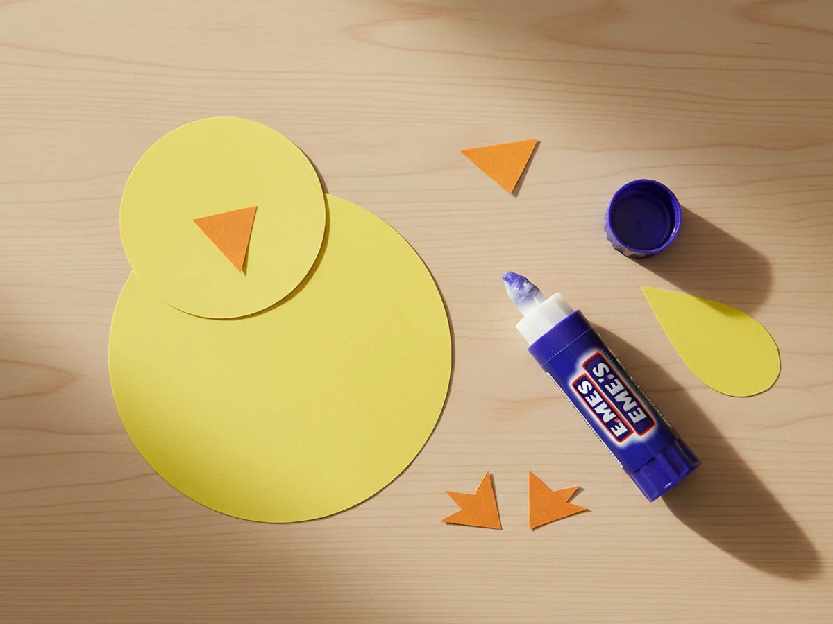 A small round yellow paper head glued onto the top of a larger round yellow body, slightly overlapping the body so the chick shape looks snuggly and connected, sitting on a white craft table with a glue stick beside it
