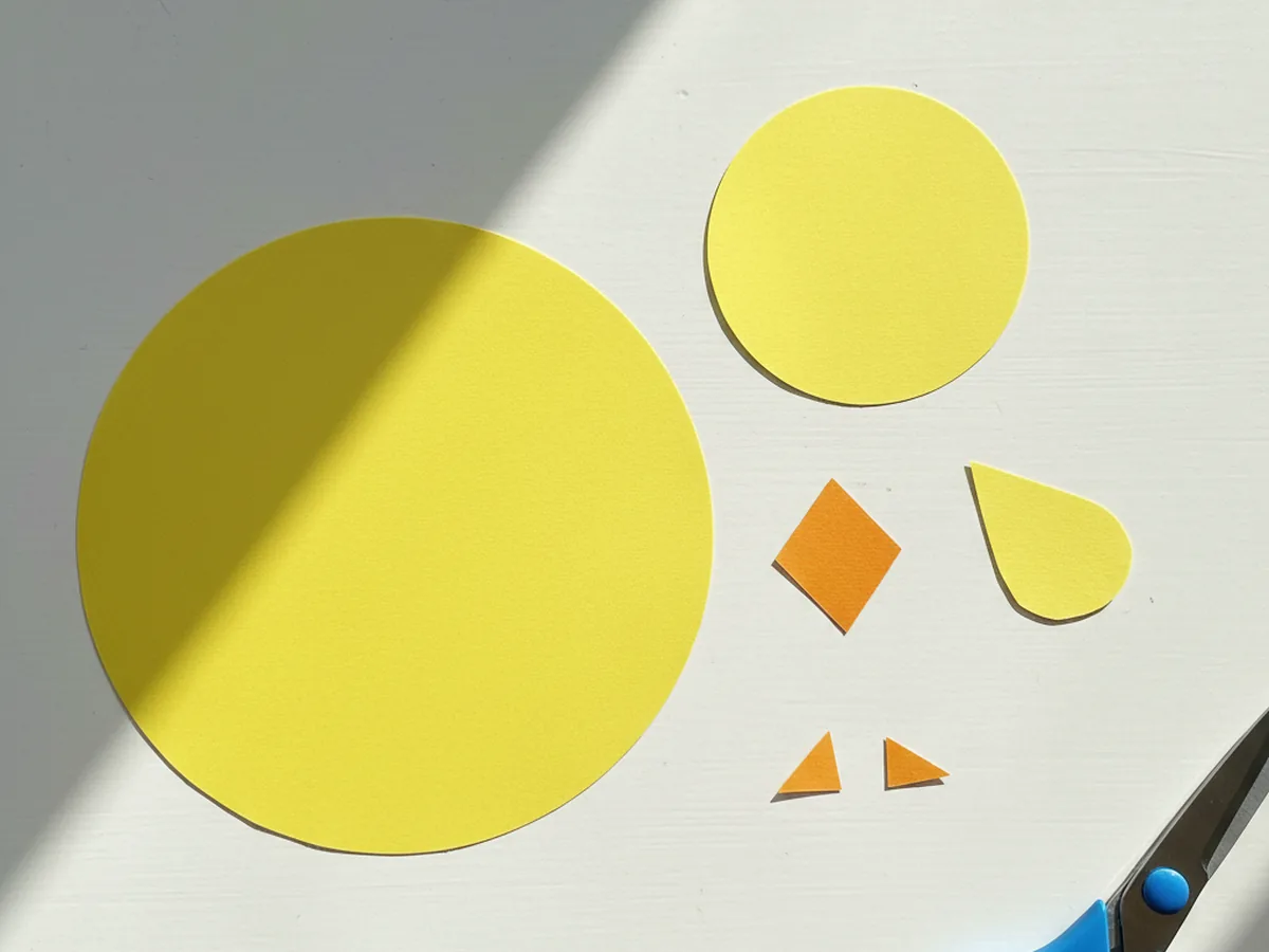 A small orange diamond beak shape, two small orange triangle feet, and a smaller yellow teardrop wing arranged neatly on a white craft table next to the larger yellow round body and head shapes for a paper chick craft