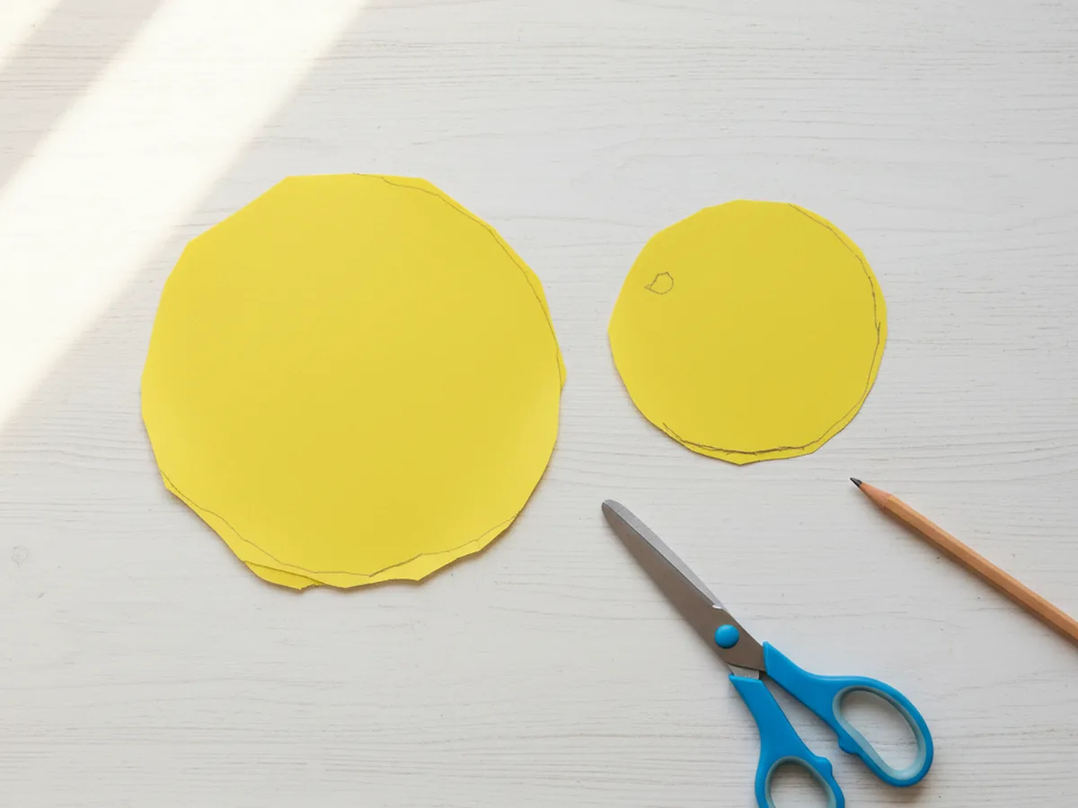 A large round body shape and a smaller round head shape cut from bright yellow construction paper resting on a white craft table next to kid scissors and a pencil, ready to be assembled into a paper chick