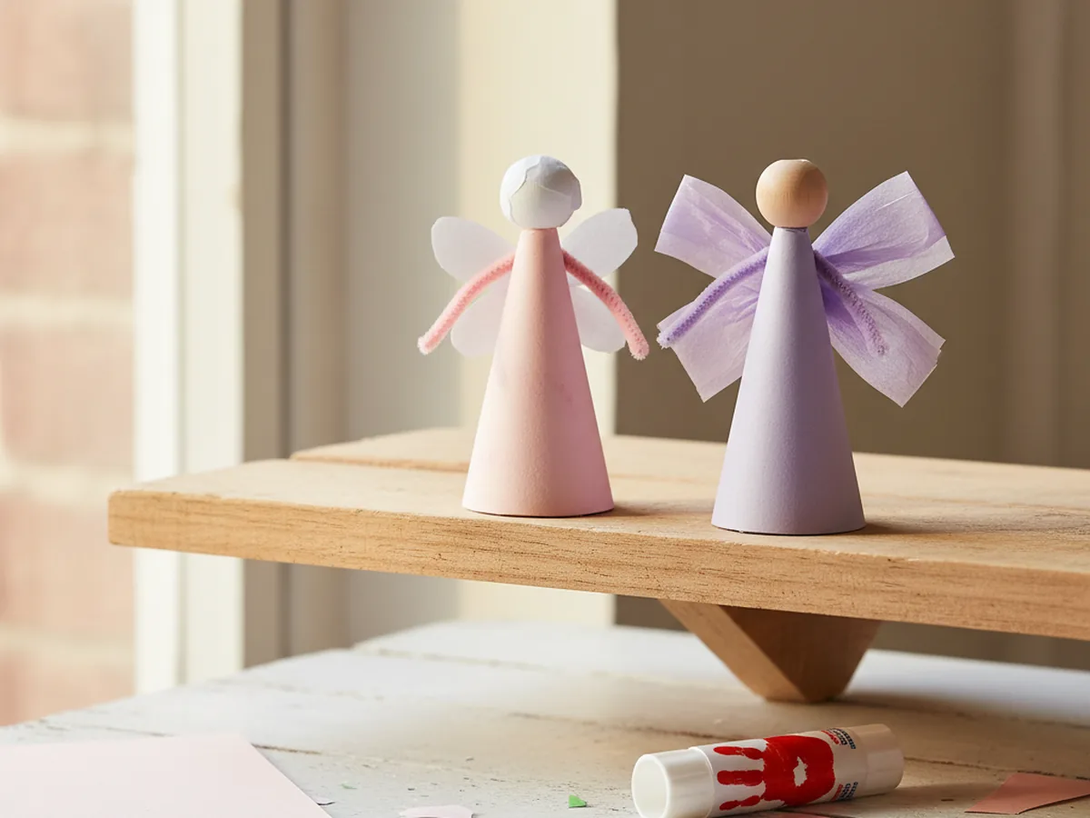 Two handmade paper cone fairy dolls in pastel pink and lavender cone dresses with paper bead heads, tiny paper wings, and pipe cleaner arms on a wooden shelf