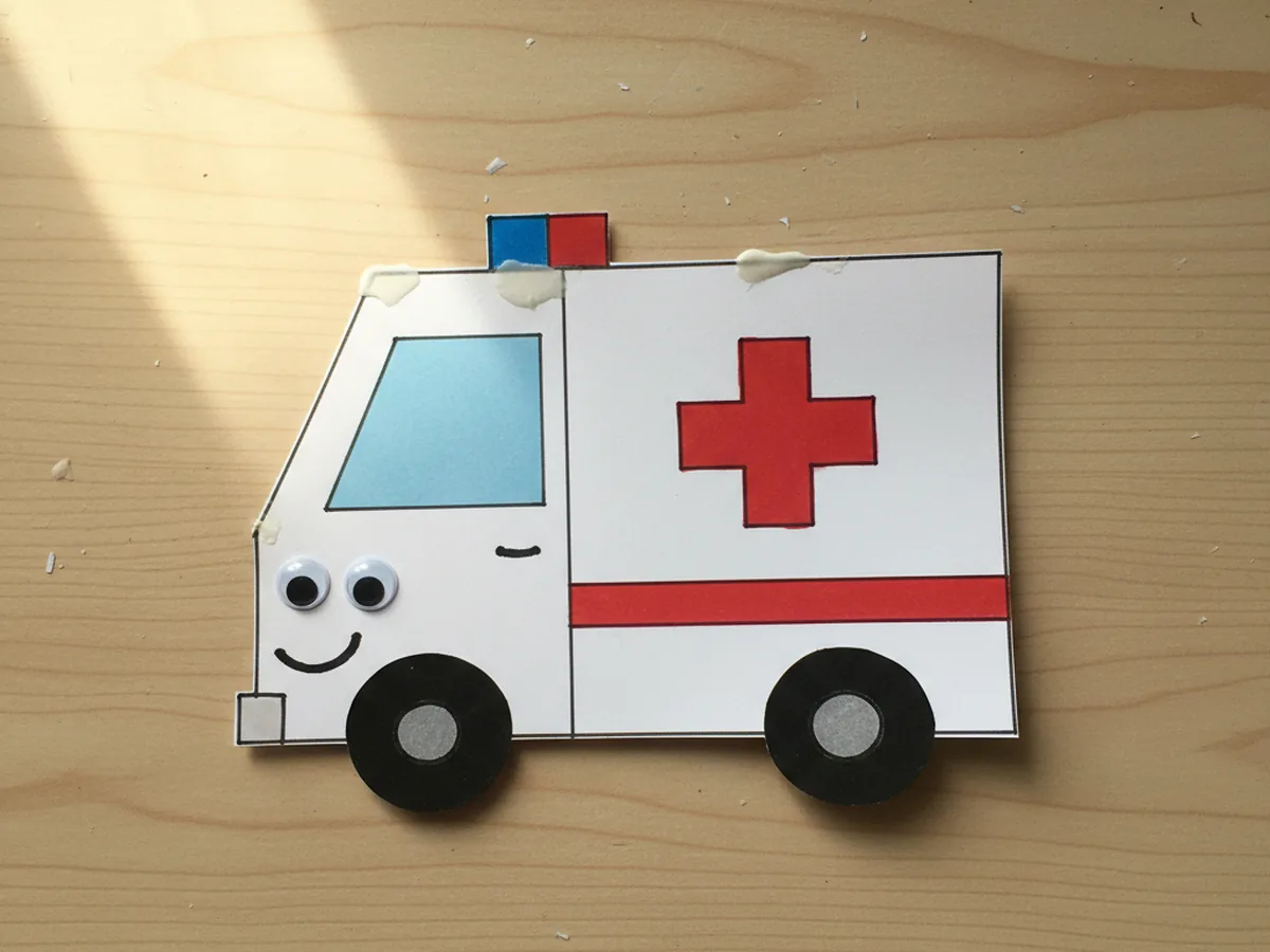 The paper ambulance with two small red and blue paper rectangles glued side by side on the roof as the siren light bar and two googly eyes and a marker drawn smile added to the front of the cab, displayed on a white craft table