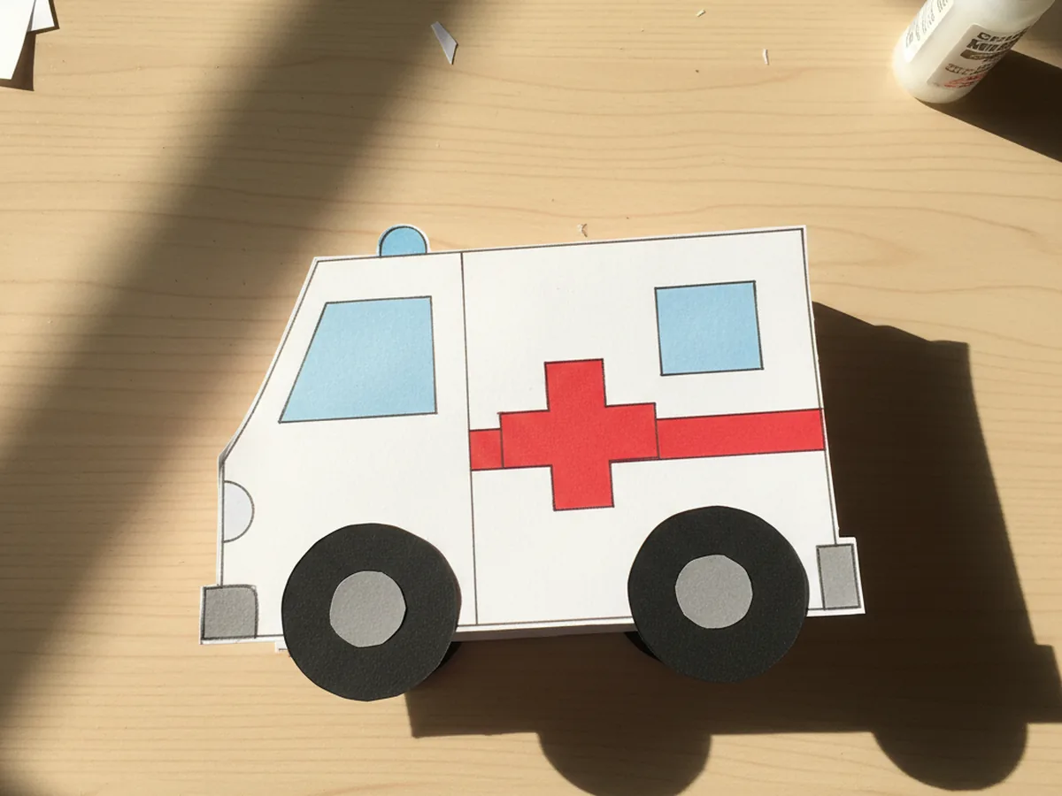 The paper ambulance now with two round black paper wheels glued under the front cab and the back box, each wheel showing a smaller grey paper circle inside as the hubcap, displayed on a white craft table