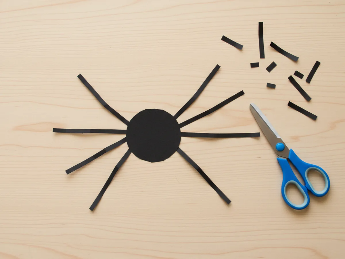 Eight thin black paper strips lined up next to a black paper circle on a craft table, ready for spider legs