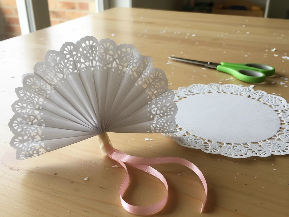 Handmade accordion-folded paper doily fan with a ribbon loop at the bottom