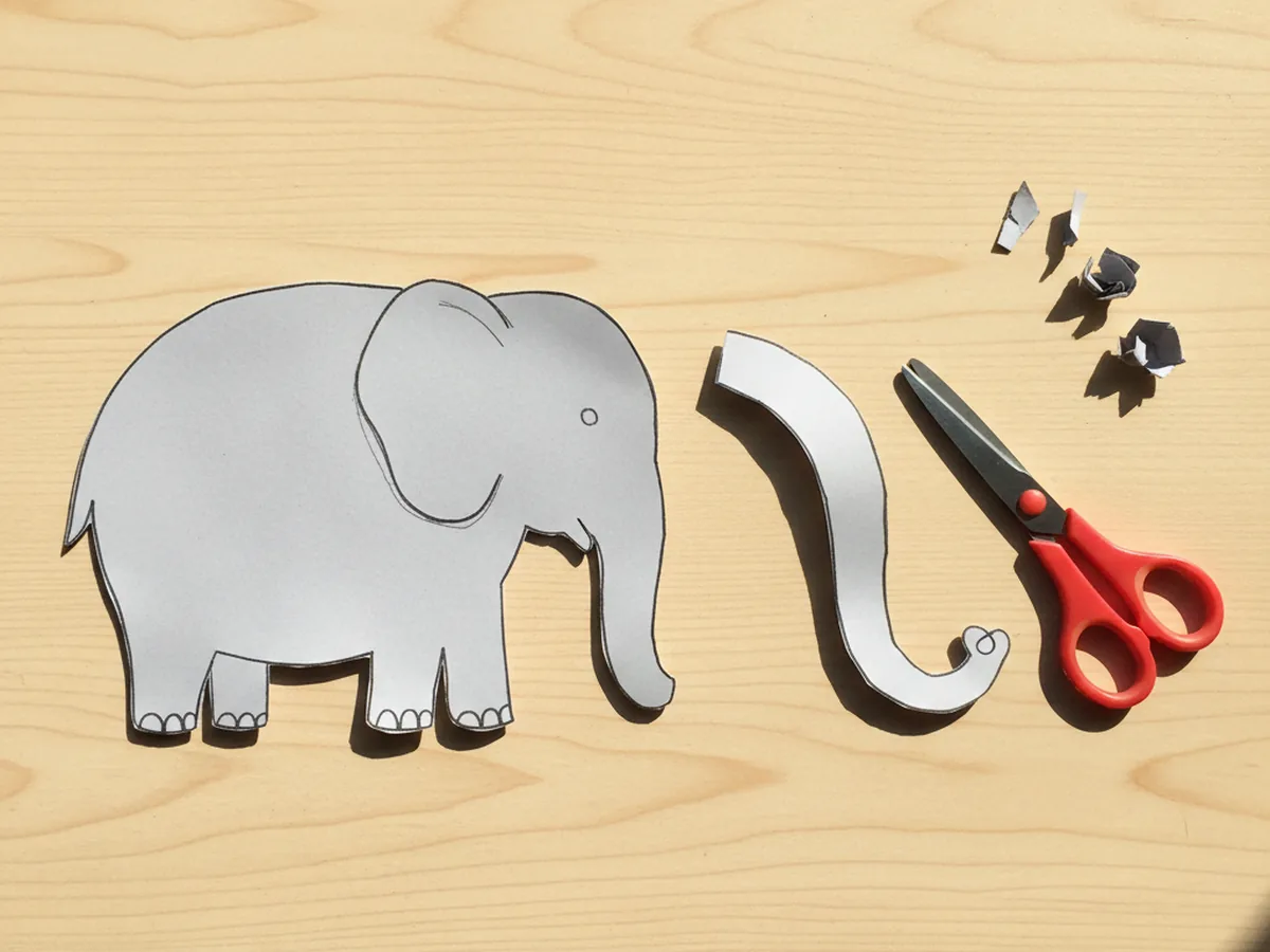 A long curved grey paper strip cut for the trunk of a paper elephant craft, lying next to the rounded grey elephant body on a craft table